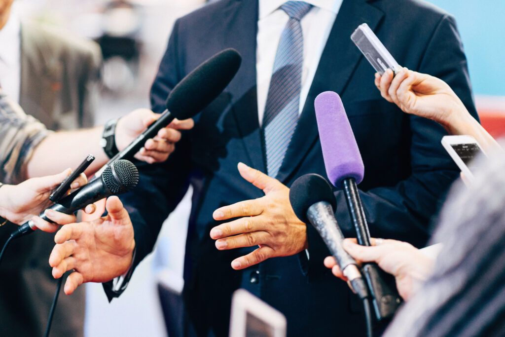 A businessman in a suit stands confidently while addressing a group of reporters holding microphones and recording devices, symbolizing media engagement and crisis communication.