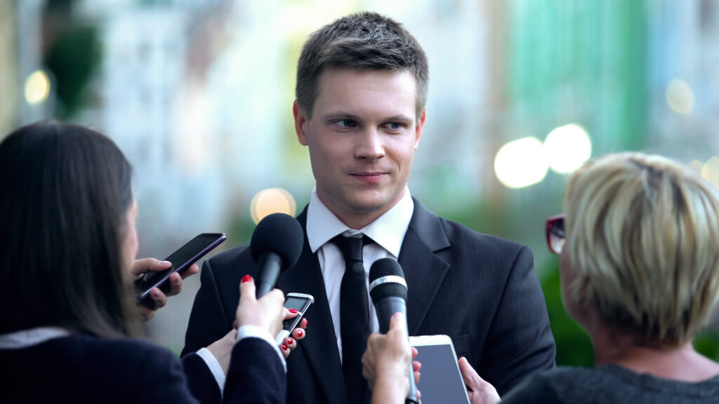 A well-dressed businessman in a black suit confidently responds to reporters holding microphones and recording devices, symbolizing media engagement and the importance of public relations in high-profile situations.