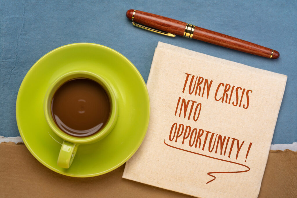 Coffee cup on green saucer next to a napkin with the handwritten phrase 'Turn Crisis Into Opportunity' and a pen, symbolizing resilience and proactive business strategy.